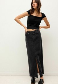Schwarzes, tailliertes Crop-Top mit quadratischem Ausschnitt, kombiniert mit einem langen schwarzen Jeansrock mit einem Schlitz vorn und rohem Saum. Schwarze Clogs.