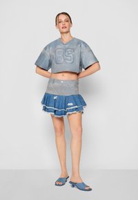 Top de mezclilla azul cortado con el número "85", combinado con una falda de mezclilla en capas con bordes deshilachados y zapatos mules azules.