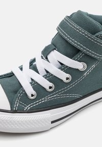 Zapatilla alta en tejido verde azulado oscuro con cordones blancos, puntera de goma y suela negra. Presenta detalles de costura y una correa de Velcro.