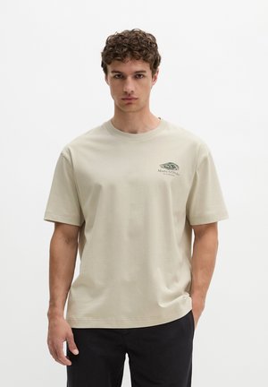 Młody mężczyzna o kręconych włosach, ubrany w beżowy t-shirt Marc O’Polo i czarne spodnie, stoi na tle jednolitej białej ściany.
