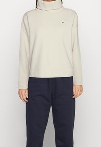 Sweter z golfem w kolorze kremowym, wykonany z miękkiego materiału, z opadającymi ramionami i żeberkowymi mankietami. Na lewej piersi znajduje się małe logo.