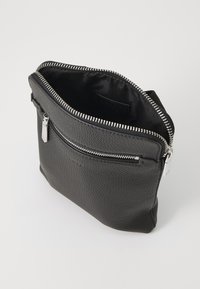 Sac en cuir noir avec un extérieur texturé, fermeture éclair argentée et une poche supplémentaire à l'avant. L'intérieur est doublé de tissu noir.