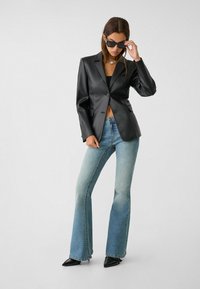 Blazer en faux cuir noir avec un seul bouton, associé à un jean évasé bleu clair et des escarpins noirs à bout pointu. Modèle portant des lunettes de soleil.