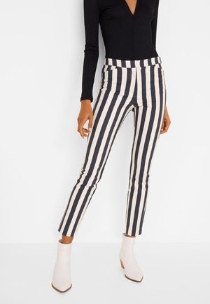 Kobieta ubrana w czarno-białe, pionowo paskowane spodnie sięgające kostek, czarną bluzkę z długim rękawem oraz białe botki, stojąca na białym tle.