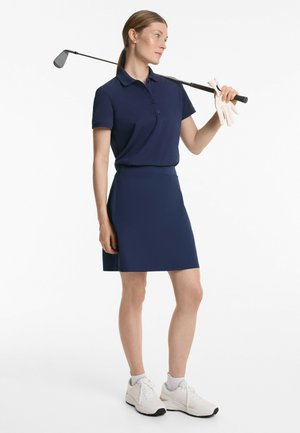 Polo marine et jupe assortie en tissu lisse ; comprend un col, des boutons et une fente sur le côté. Des baskets blanches complètent la tenue.