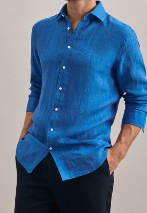 Hombre con camisa de lino azul de manga larga con botones blancos, manos en los bolsillos de pantalones negros, sobre un fondo liso.