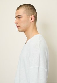 Baltas megztas V formos džemperis su laisvu siluetu, minkšta tekstūra ir nuleistais pečiais, demonstruojamas ant modelio iš šono, prieš neutralų foną.