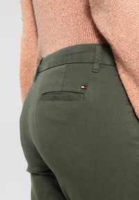 Olijfgroene broeken met een achterzak met een klein rood, wit en blauw logoaccent. Gemêleerde lichtroze trui zichtbaar erboven.