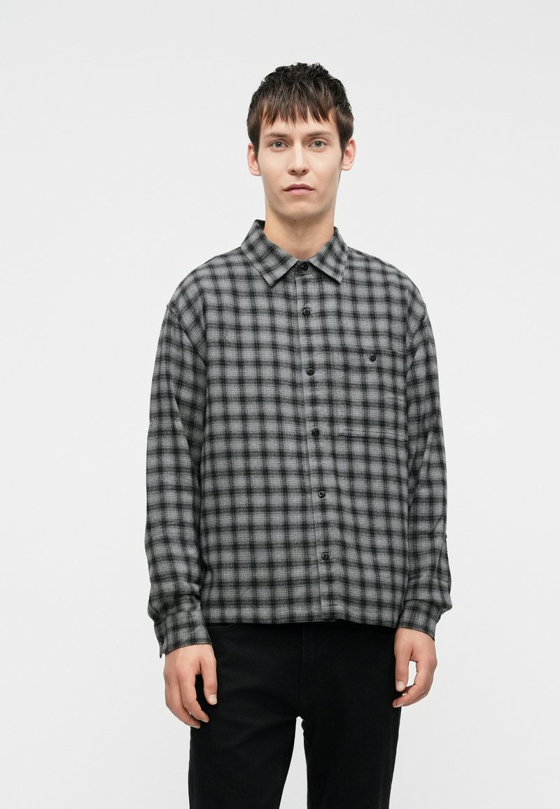 Šedá a černá kostkovaná košile, design na knoflíky, dlouhé rukávy, náprsní kapsa, tkaná struktura látky, volný střih. Model stojí na bílém pozadí.