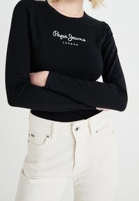 Person wearing a black long-sleeve top with "Pepe Jeans London" logo and high-waisted off-white corduroy pants, arms crossed.
