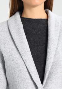 Blazer texturizado gris claro con un amplio cuello de chal sobre una blusa de punto gris oscuro, mostrando materiales suaves y un look en capas.