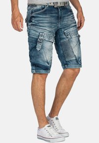 Pantaloni cargo in denim di colore azzurro chiaro con diverse tasche, dettagli testuali sulle gambe e un aspetto leggermente sbiadito. Indossati con sneakers bianche.