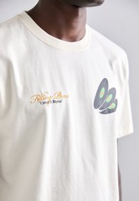 Jasno beżowa bawełniana koszulka z krótkim rękawem, z złotym haftem z napisem "Filling Pieces" oraz grafiką trzech czarnych winylowych płyt z zielonymi akcentami.