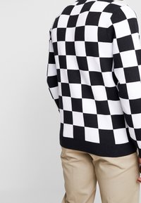 Pull à damier noir et blanc avec un ourlet et des poignets côtelés, arborant un design classique à col rond. Le matériau semble tricoté et doux.