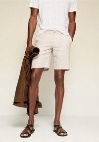 Helle beige Leinen-Shorts mit slim Fit und Knopfverschluss. Kombiniert mit einem grauen T-Shirt und einer braunen Wildlederjacke, ergänzt durch schwarze Sandalen.