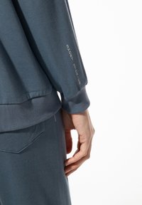Blauwe katoenen sweatshirt met een gestructureerde stof en geborduurde "SHORT STORIES" detail op de mouw. Ontspannen pasvorm met geribde manchetten.