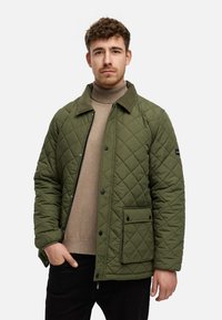 Quiltet olivengrøn jakke med en kordfløjlskrave, lomme foran og trykknapper, båret over en beige turtleneck og sorte bukser.