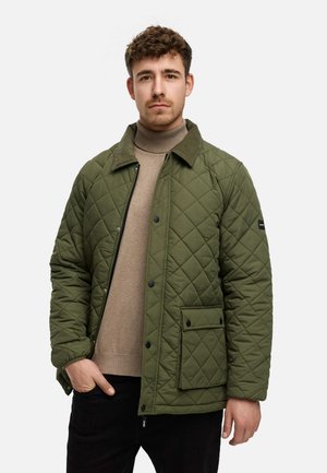 Chaqueta acolchada de color verde oliva con cuello de pana, bolsillo frontal y botones a presión, llevada sobre un jersey de cuello alto beige y pantalones negros.
