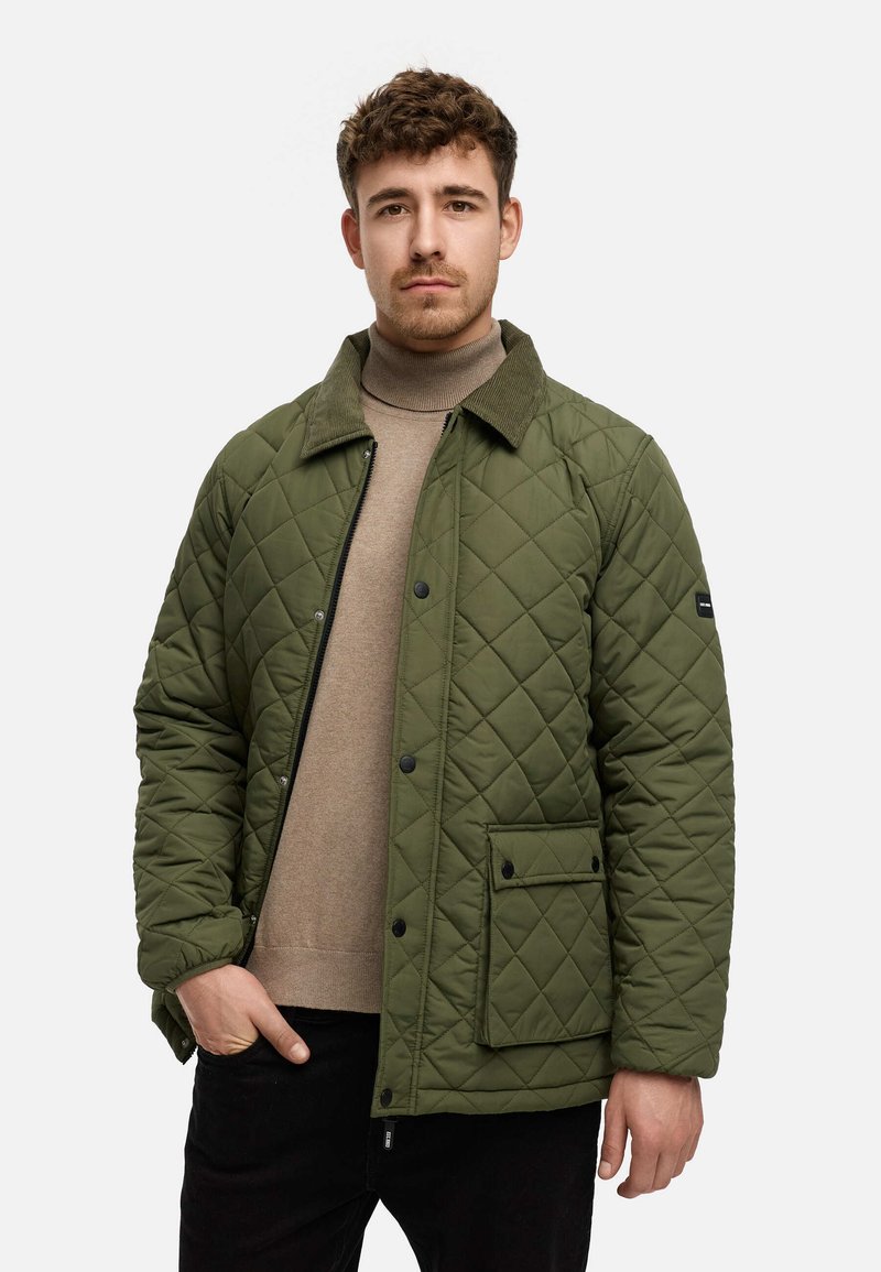 Quiltet olivengrøn jakke med en kordfløjlskrave, lomme foran og trykknapper, båret over en beige turtleneck og sorte bukser.