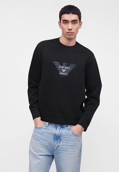 Νέος άνδρας που φοράει μαύρο φούτερ Emporio Armani και ανοιχτό μπλε τζιν, στέκεται με τα χέρια στις τσέπες σε ένα απλό φόντο.