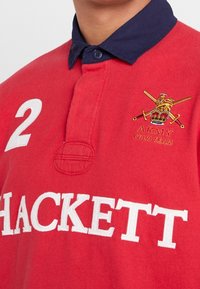 Polo rouge avec un col marine, arborant le texte brodé blanc "HACKETT" et un logo doré avec des épées croisées et un emblème de lion.