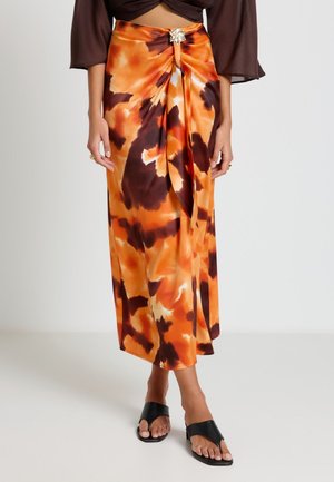 Femme portant une jupe midi tie-dye orange et marron avec un nœud devant et une broche feuille argentée décorative, associée à des sandales noires ouvertes aux orteils.