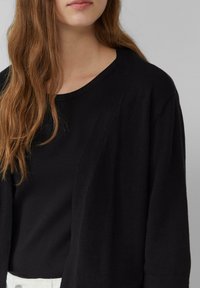 Zwarte cardigan met een relaxte pasvorm, gemaakt van zachte stof. Heeft een diepe V-halslijn en een open voorkant, gelaagd over een zwart top.