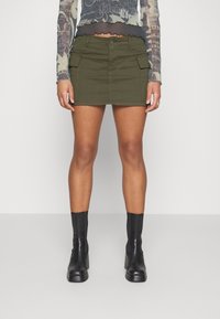 Nainen, jolla on päällä oliivinvihreä cargo-minihelma, mustat nilkkurit ja läpikuultava, kuviollinen pitkähihaiset paita, seisoo tasaisella taustalla.