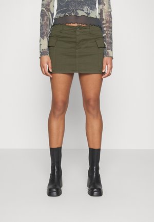 Vrouw in een olijfgroene cargominirok, zwarte enkellaarzen en een doorschijnend, geprint langemouwen-top, staand tegen een effen achtergrond.