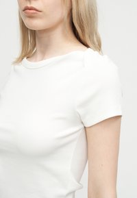 Νεαρή γυναίκα με ανοιχτό ξανθό μαλλί, που φοράει μονόχρωμο λευκό κοντομάνικο μπλουζάκι, σε ουδέτερο φόντο, η εικόνα κομμένη στο πηγούνι και στον άνω βραχίονα.