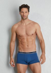 Modré boxerky s těsným střihem, vyrobené z měkké tkaniny. Mají kontrastní opasek s logem a hladkou texturu.