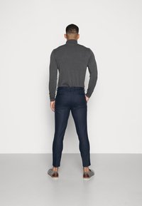 Muž oblečený v tmavě šedém roláku, námořnických slim-fit kalhotách a šedých slip-on botách, stojí proti bílé zdi s obráceným zády.