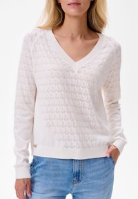 Lichtbeige gebreide trui met een V-hals, een gestructureerd hexagonaal patroon, ribbelmanchetten en een ribbelzoom. Draagt met blauwe jeans.