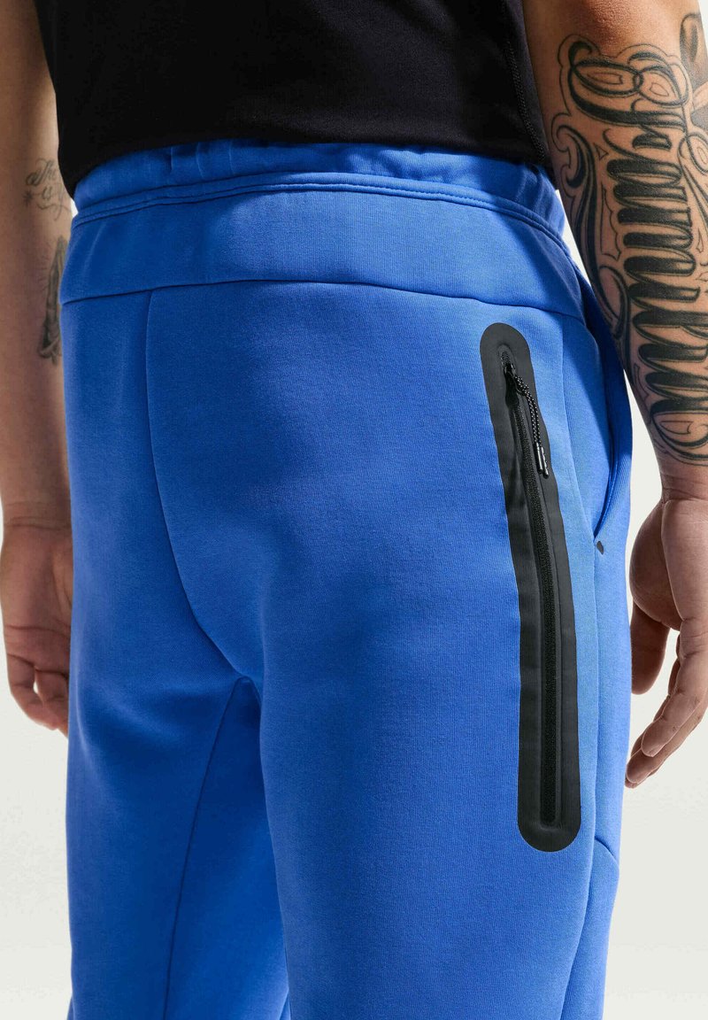 Personne portant un pantalon bleu vif avec une poche verticale noire zippée sur la cuisse et des tatouages visibles sur le bras, debout devant un fond uni.