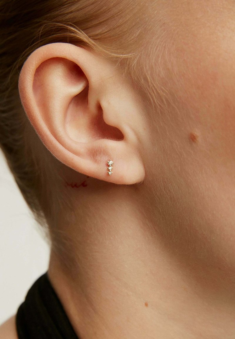 Pendiente de oro con tres pequeñas piedras preciosas transparentes, con un diseño elegante y minimalista, exhibido en primer plano cerca de una oreja.