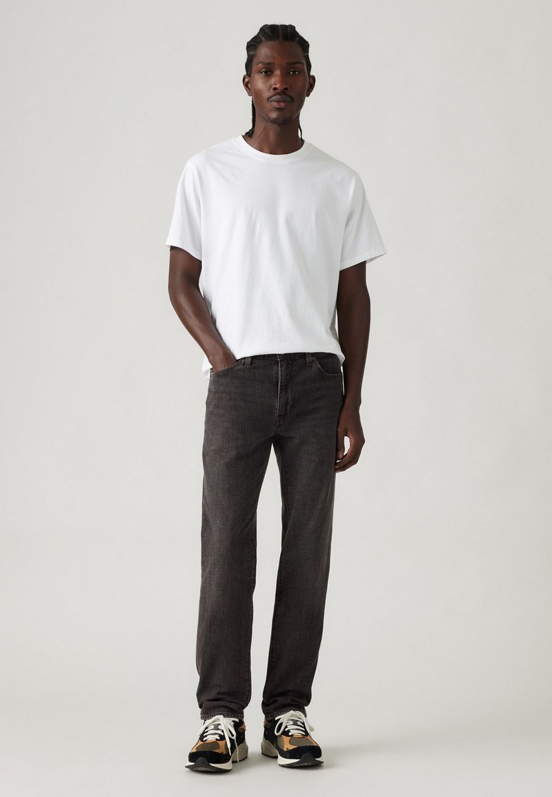 Camiseta blanca de algodón combinada con pantalones de mezclilla gris oscuro. El modelo lleva zapatillas negras y beige, presentando un atuendo casual en tonos neutros.