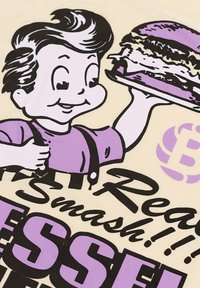 Camiseta beige con un personaje de dibujos animados de cabello oscuro, sosteniendo un sándwich. El texto en negrita negro y púrpura dice "¡Real Smash!!!"