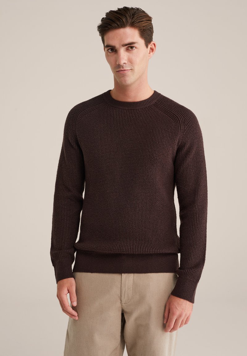 Tumši brūns teksturēts adījums ar apaļu kakla izgriezumu un ribotām piedurknēm, saskaņots ar gaiši bēžām biksēm.