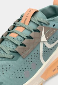 Nike trailrun schoen met een teal mesh bovenwerk, perzikaccenten, rubberen zool, gestructureerde grijze overlays en reflecterende details.