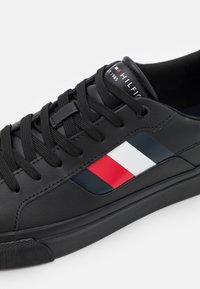 Zapatilla de cuero negra con cordones negros, que presenta un diseño lateral de rayas rojas, blancas y azules. Suela de goma texturizada y marca en la lengüeta.