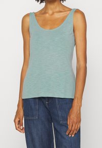 Ljus teal färgad linne gjord av mjukt tyg, med en rundad halsringning och tunna axelremmar. Slät textur utan ytterligare mönster.