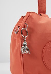 Pomarańczowa torba materiałowa z okrągłym naszywką logo i srebrnym metalowym breloczkiem w kształcie robota przymocowanym do pierścienia na pasku bocznym na białym tle.