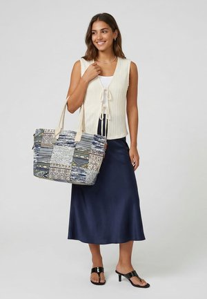 Femme souriante, portant un haut en maille crème sans manches, une jupe en satin bleu marine, des sandales à talons noires, portant un grand sac cabas à motifs sur l’épaule.