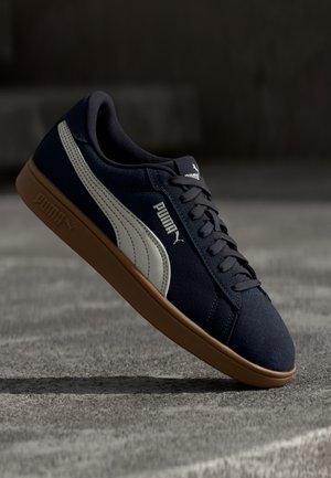 Granatowy sneaker Puma z białym paskiem i gumową podeszwą, unoszący się nad szorstką betonową powierzchnią w miękkim oświetleniu.