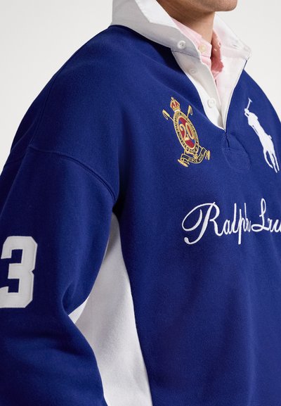 Muž ve modro-bílém rugby tričku Ralph Lauren s vyšitým erbem, číslem 3 na rukávu a pod ním růžovým košilovým límečkem.