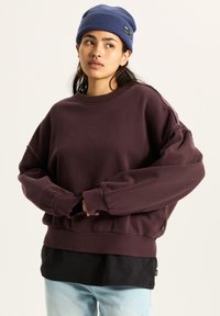 Felpa bordeaux con spalle scese, polsini e orlo a coste, sovrapposta a una maglietta nera. Indossata con un berretto blu. Tessuto morbido.