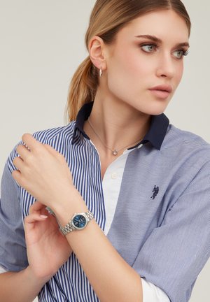 Giovane donna con capelli castano chiaro raccolti in una coda di cavallo, che indossa un orologio d'argento, piccoli orecchini a cerchio, una collana delicata e una camicia a righe con bottoni.