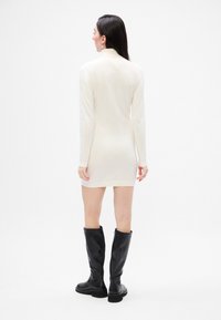 Rochie albă cu mâneci lungi și guler înalt, textură netedă și croială ajustată; asociată cu cizme negre până la genunchi, cu un design elegant.