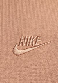 Nike logo izšūts ar saskaņotu diegu uz gaiši brūna adīta auduma fona.
