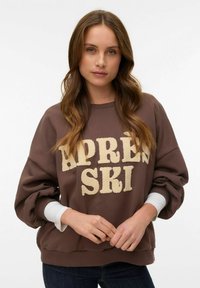 Sudadera marrón con las palabras "APRES SKI" en letras beige texturizadas. Presenta mangas anchas y un dobladillo acanalado. Se lleva con una camiseta blanca de manga larga debajo.